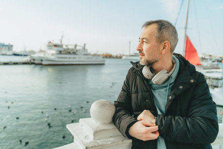 A middle-aged man in a black jacket and full-size headphones in front of yachts.の写真素材