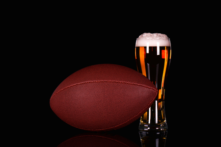 Beer glass with dark beer foam and American football ball on black background.の写真素材