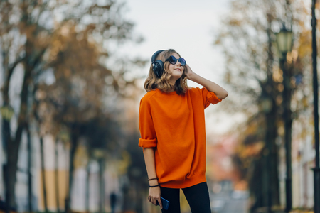 cute young girl walking down old city street and listening music in headphones, urban style, stylish hipster teen in black hat listen music and smile, orange crazy styleの写真素材