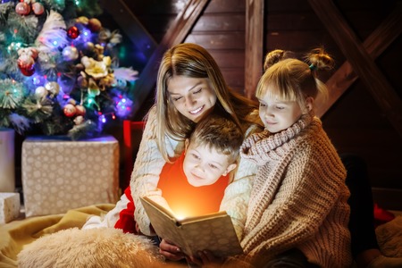 Merry Christmas and Happy New Year. Beautiful family in Xmas interior. Pretty young mother reading a book to her daughter and son near Christmas tree.の写真素材