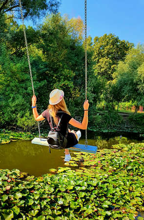 Beautiful young blonde woman with a hat on her head, riding in a swing over the water lily lake....