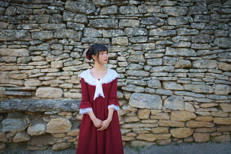 Young Chinese woman in red retro style dress standing in front of a brick wall of an ancient French...