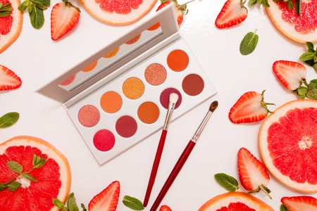 Cute flat lay with palette of cosmetics with fresh fruit, cut strawberries and grapefruit or red orange, mint leaves on a white background.の写真素材
