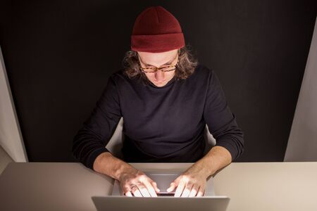 Designer or programmer in the workplace. young man in a hat and glasses sitting at a laptop. dark...