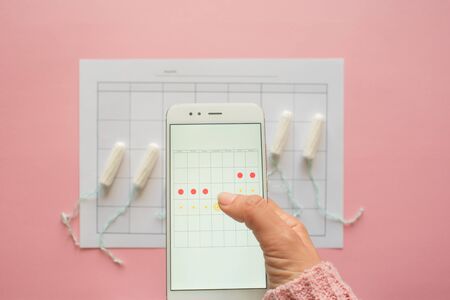 Menstrual cycle. Calendar for the month with marks and a mobile application on the smartphone screen. Phone in hand on pink backgroundの写真素材