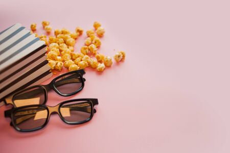 Two black 3d glasses lie next to a black and white papper striped box of popcorn, scattered on a pink background.の写真素材