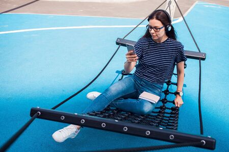 The girl, a brunette, pretty, with glasses, sits in a hammock, sways, on the playground with a bright blue coating, listening to music, dancing, relaxing, singing, trying to catch a wifi. Outdoor.の写真素材