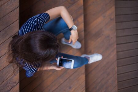 A brunette girl, a student with long hair, with a large wrist watch, in jeans, sits on wooden steps and checks a smartphone. The view is clearly from above. Outdoor.の写真素材