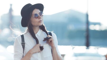 Young woman in hat and round sunglasses walking in city. Girl tourist enjoys the walk. Hipster in a hat with a backpack, funny portraitの写真素材