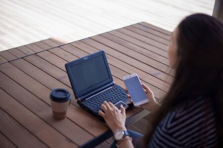 A brunette girl with a large wrist watch, half-turned from the back, works at a laptop standing on a wooden table. Nearby is a smartphone. Checks gadget. Rear view, side, partially. Outdoor.の写真素材