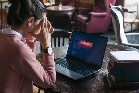 Something went wrong. A very excited girl in a pink sweater sits at an old desk in the library, in front of her is a laptop, on the display of which the site code is closed with a red error warning.の写真素材
