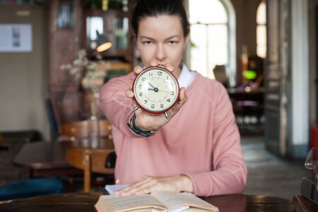 Deadline. A closeup alarm clock aimed at the frame held by a brunette girl in a pink sweater sitting...