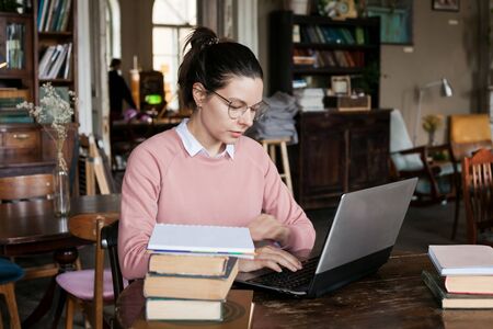 Exam preparation. Attractive brunette student girl in glasses and pink sweater, sits at a laptop, is...