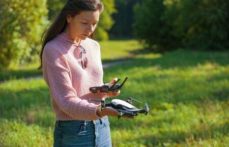 A young woman in a clearing in a park launches, checks, holds a drone, in one hand, in the other...