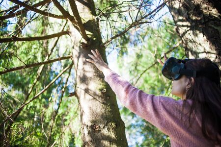 Virtual forest. A young woman among the trees, wearing virtual reality glasses, touches the boles and branches, the air, on a sunny summer day, straightens her helmet. Side and back view. Copyspace.の写真素材