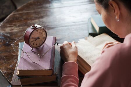 Too little time. An alarm clock stands on a stack of books, next to glasses, on a table in an old library, in front of a girl bent over a book. Stress during exams. Top view and over the shoulder.の写真素材