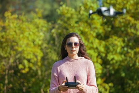 The four-propeller drone hovered in the air at eye level. In the background, a girl in sunglasses and a pink sweater is watching over his flight with control panel in hands. The foreground is blurred.の写真素材