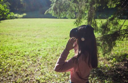 Virtual forest. A young woman in virtual reality glasses stands under the trees in a clearing, on a sunny summer day, touches the air with her hands, straightens her helmet. Side view. Copyspace.の写真素材
