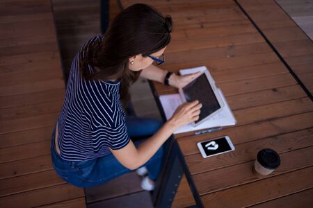 A serious brunette girl, a freelancer, with a large wrist watch, works at a wooden table on a...