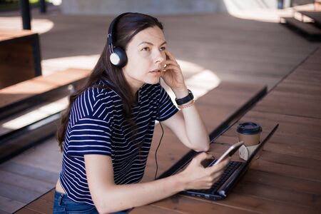 girl student with a smartphone, sits at a laptop and listens carefully in headphones, learns foreign...