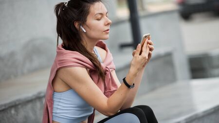 Portrait of a sporty young woman in sports uniform sitting, resting on the steps with a smartphone and wireless headphones after a warm-up, exercises. Side view. Copyspace.の写真素材