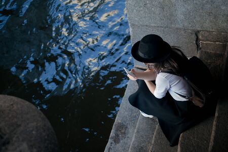 young woman in a hat and with a backpack walks in the city and uses a smartphone. Hipster on a walk uses the phone and takes photos for social networksの写真素材
