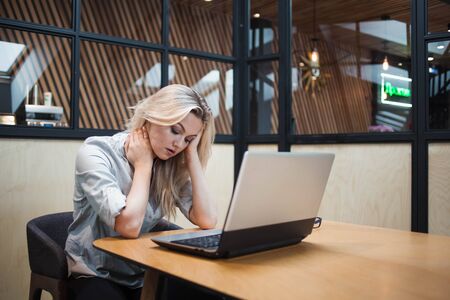 Tired overworked young woman in the office at the laptop. Office worker at the table, sitting clutching headの写真素材