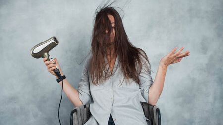 Hair dryer, an attempt to do styling for long and unruly hair. Hairstyle styling and hair care, portrait on a gray backgroundの写真素材