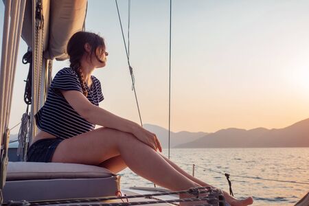 attractive young woman in a striped t-shirt enjoys the sunset on the deck of a sailing yacht. Girl yachtsman looks into the distance. Sailing regatta, sea voyageの写真素材