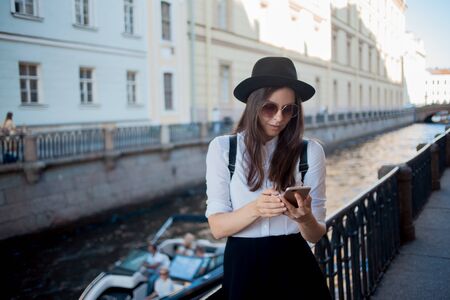 young woman in a hat and with a backpack walks in the city and uses a smartphone. Hipster on a walk uses the phone and takes photos for social networksの写真素材