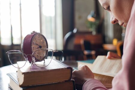 Too little time. An alarm clock stands on a stack of books, next to glasses, on a table in an old library, in front of a girl bent over a book. Stress during exams. Top view and over the shoulder.の写真素材