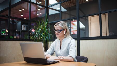 young blonde woman sits at her laptop and uses wireless headphones. Freelancer or student uses...