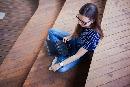 A brunette girl, a student with long hair, with a large wristwatch, with glasses sitting cross-legged on wooden steps and typing on a laptop lying on her lap, works as freelancer. Top view. Outdoor.の写真素材