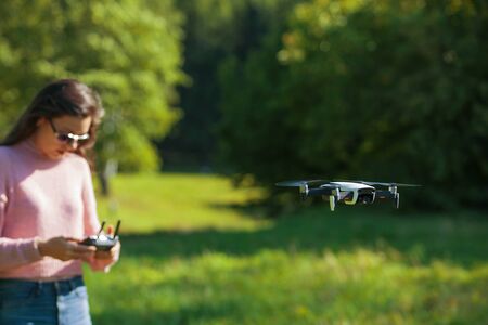 The four-propeller drone hovered in the air at eye level. In the background, a girl in sunglasses and a pink sweater is watching over his flight with control panel in hands. The foreground is blurred.の写真素材