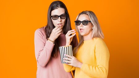 Two friends watch an interesting movie and eat popcorn. Two young female moviegoers, concept on yellow backgroundの写真素材