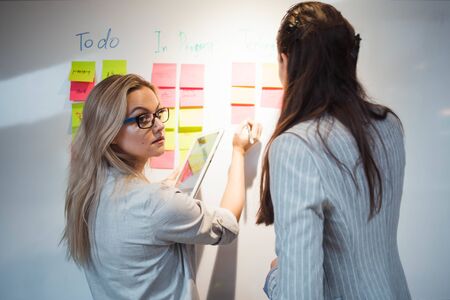 Teamwork, project management, agile methodology. Two young business women in the office are working on a startup. Colleagues plan work and stages of development of company, glue sticky leaves on Boardの写真素材