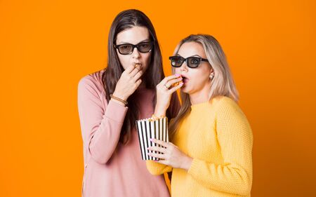 Two friends watch a movie or TV series and eat popcorn. Two young women fans of the movie, the concept on yellow backgroundの写真素材