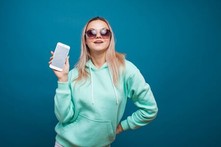 Stylish trendy hipster girl in the blue hoodie with the hood uses a smartphone. Happy young blonde...