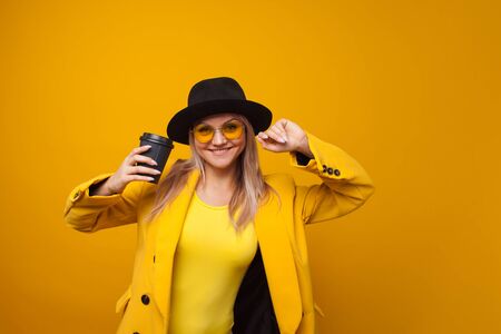 Take-away coffee. Stylish trendy young woman in bright clothes on a yellow background. A cool blonde girl in a yellow jacket, yellow sunglasses and a black hat drinks coffee and is happyの写真素材
