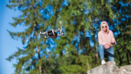 A young woman in a pink hoodie and sunglasses sitting on a stone bench and operated by remote control drone flight in nature. Full-length photo. Blurred background. Drone in the foreground.の写真素材