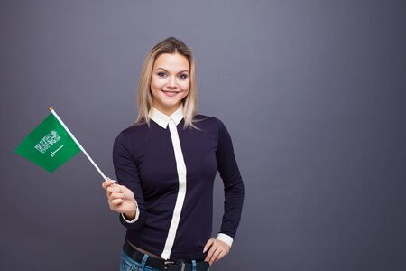Immigration and the study of foreign languages, concept. A young smiling woman with a Saudi Arabia flag in her hand. Girl waving a Saudi Arabia flag on a gray backgroundの写真素材