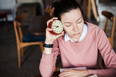 Lack of sleep. A student girl, sitting with an alarm clock on her temple and closing her eyes, counts short minutes of break. Affects stress and lack of sleep. Stress during exams.の写真素材