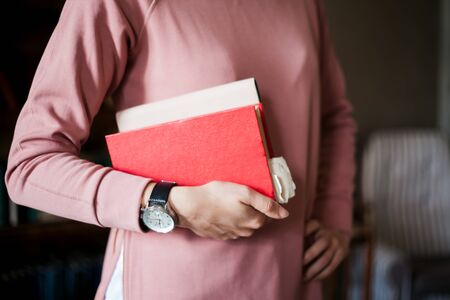 A girl in a pink sweater and with large clock holds armpit in a stack of books, against the background of the interior of an old library, bookshelf and houseplants. The background is slightly blurred.の写真素材