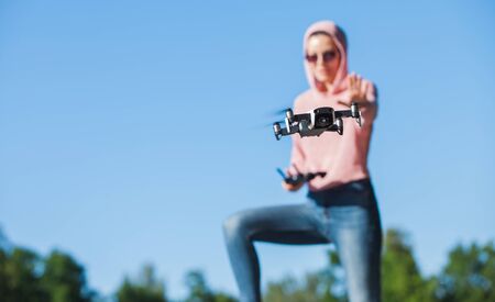 Standing with a bended knee, a young woman in pink hoodie and dark glasses controls the drone's outdoor flight control panel in nature. Full-length photo. Blurred background. Drone in the foreground.の写真素材