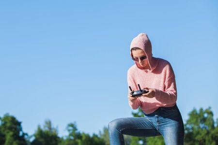 Young woman wearing sunglasses and hoodie holding a remote control, controls, controls are not visible in the frame drone. A woman carefully looks into the distance against a blue sky. Copyspace.の写真素材
