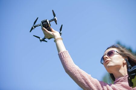 A young woman in a clearing in a park launches, checks, holds the drone with her outstretched arm above her head, in another control panel. Outdor. Copyspace. The woman is partially visible.の写真素材