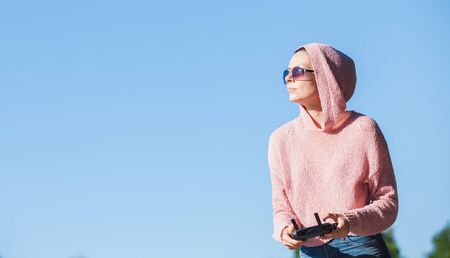 Young woman wearing sunglasses and hoodie holding a remote control, controls, controls are not visible in the frame drone. A woman carefully looks into the distance against a blue sky. Copyspace.の写真素材