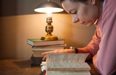 Diligent student. A tired girl student leaned over a thick book with many bookmarks, at a tiny...