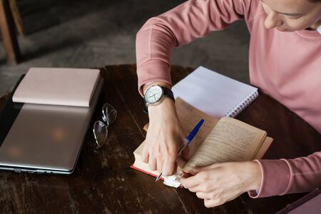 A girl in a pink sweater leafs through a thick book with many bookmarks, makes notes, nibbles a pen,...