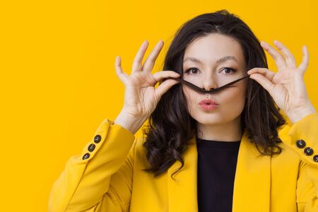 Stylish young woman in a yellow jacket on a yellow background, making a mustache out of her hair....